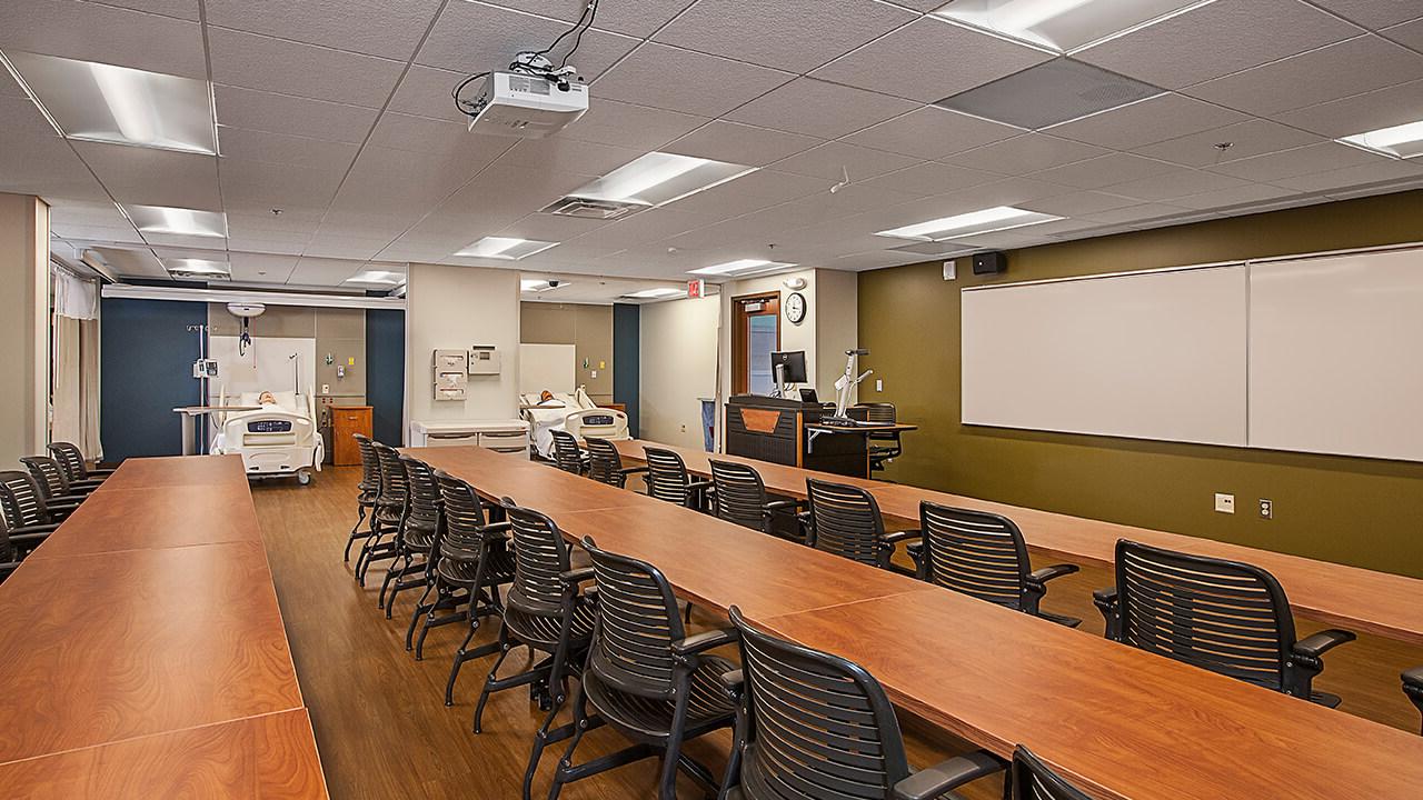 A smaller classroom with three stretches of tables, each lined with seats. At the front of the room is a teaching station with a projector and whiteboards. On the far side are two simulation hospital rooms; open to the classroom, including beds and manikins.